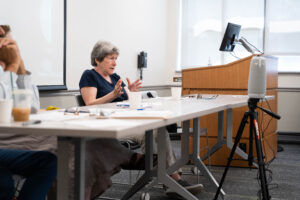 nancy wackstein serving as one of the day's panelists. she is sitting behind a white table, wearing a dark blue shirt.