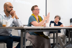 melissa aase speaks to the audience. she is wearing a turquoise shirt with a yellow scarf wrapped around her neck.