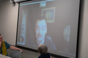 tony armstrong appears over the projector screen via zoom and speaks to the audience. he is wearing a blue shirt.