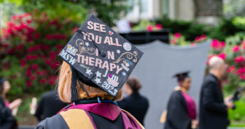 a student dressed in a cap and gown for graduation. the back of their graduation cap reads "see you all in therapy."