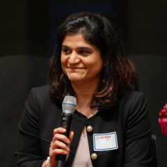 supriya chabria speaks on a panel. she is holding a microphone and smiling.