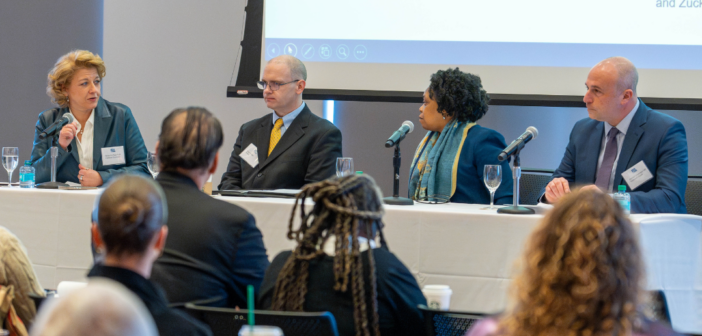McPhee and her panel sit at a long table on a stage, engaging in a discussion. The audience is visible in the foreground, while a projector screen is displayed in the background.