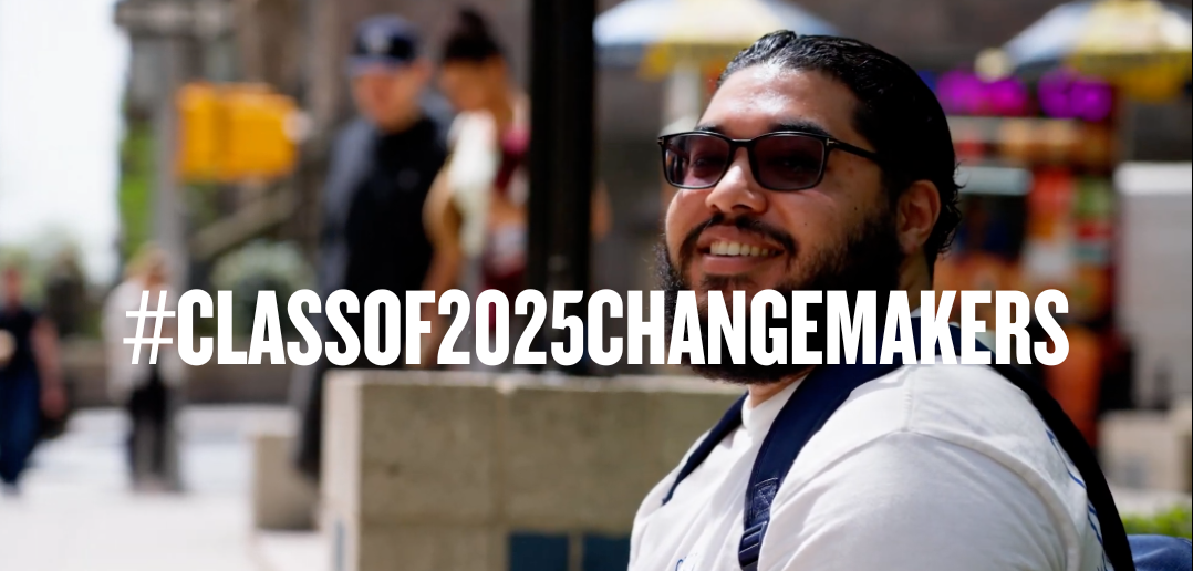 Giancarlos Valerio sits outside the Fordham University Lincoln Center campus, smiling. Blurred pedestrians and colorful umbrellas appear in the background. Bold white text across the image reads “#CLASSOF2025CHANGEMAKERS.”